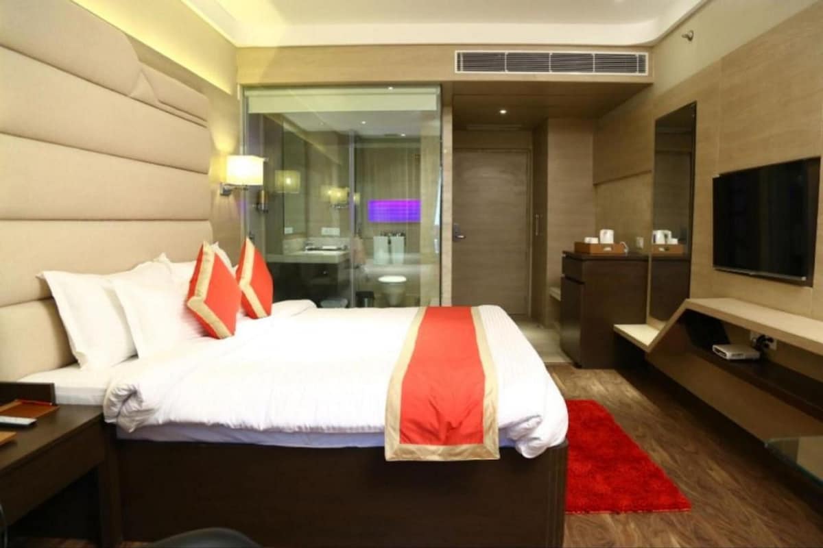 A modern hotel room with a large bed, a TV, a desk, and a bathroom visible through a glass partition.