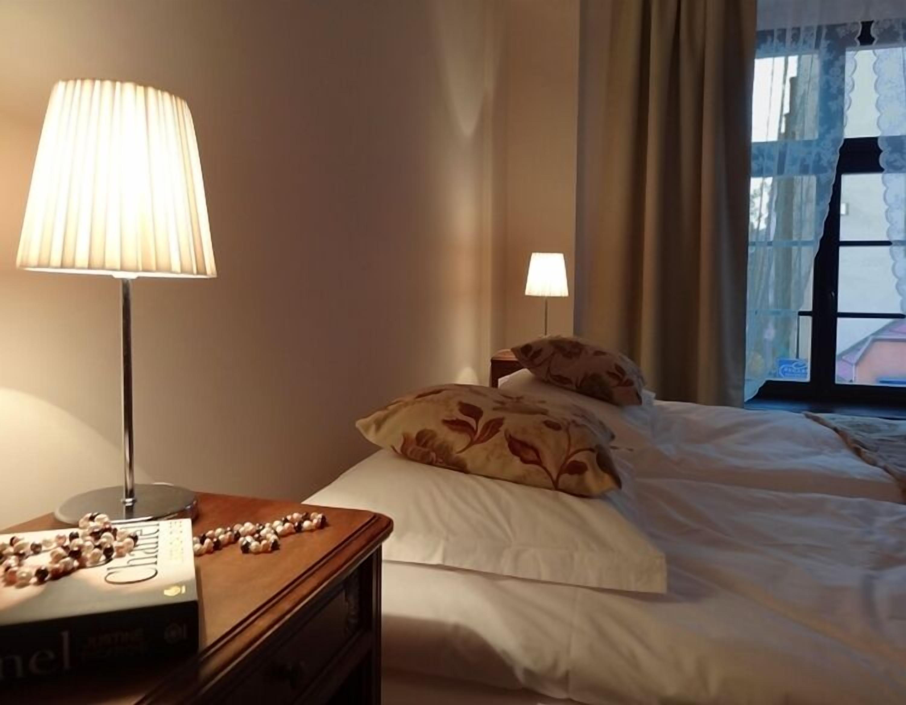 A hotel room with a bed, a bedside lamp, a nightstand with a book and decorative beads, and a window with curtains.