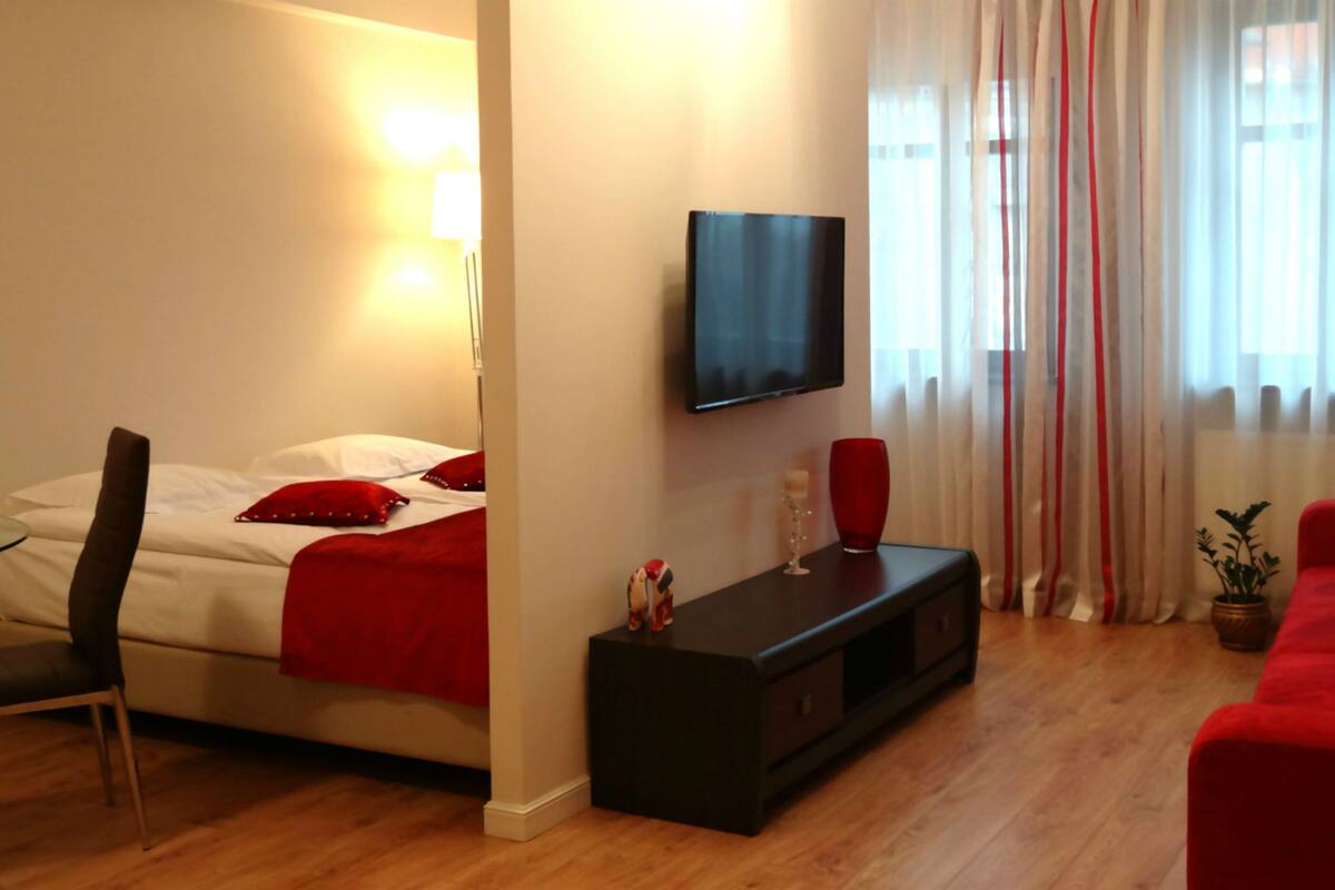 Een moderne woonkamer met een bed, een aan de muur bevestigde televisie, een rode bank en een zwarte tv-meubel.
