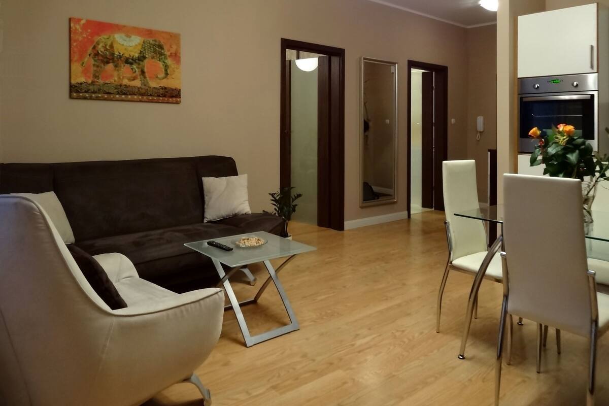 Een woonkamer met een donkerbruine bank, een glazen salontafel en een eethoek met witte stoelen en een glazen tafel.