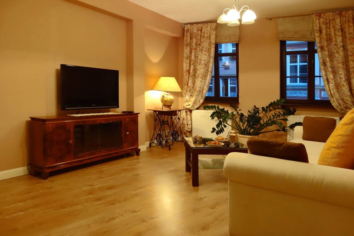 Een woonkamer met een flatscreen televisie op een houten kast, een salontafel met een lamp en decoratieve objecten, een bank met kussens, en een raam met gordijnen.
