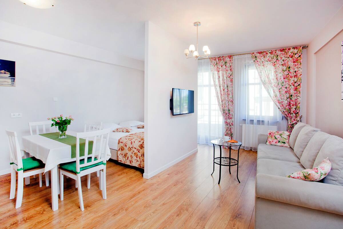 Een moderne woonkamer met een bank, een eettafel met stoelen, een aan de muur bevestigde televisie en een raam met bloemen gordijnen.