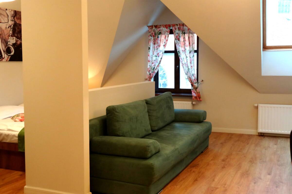 Een kamer met een groene bank, een raam met bloemen gordijnen, een radiator en een televisie aan de muur.