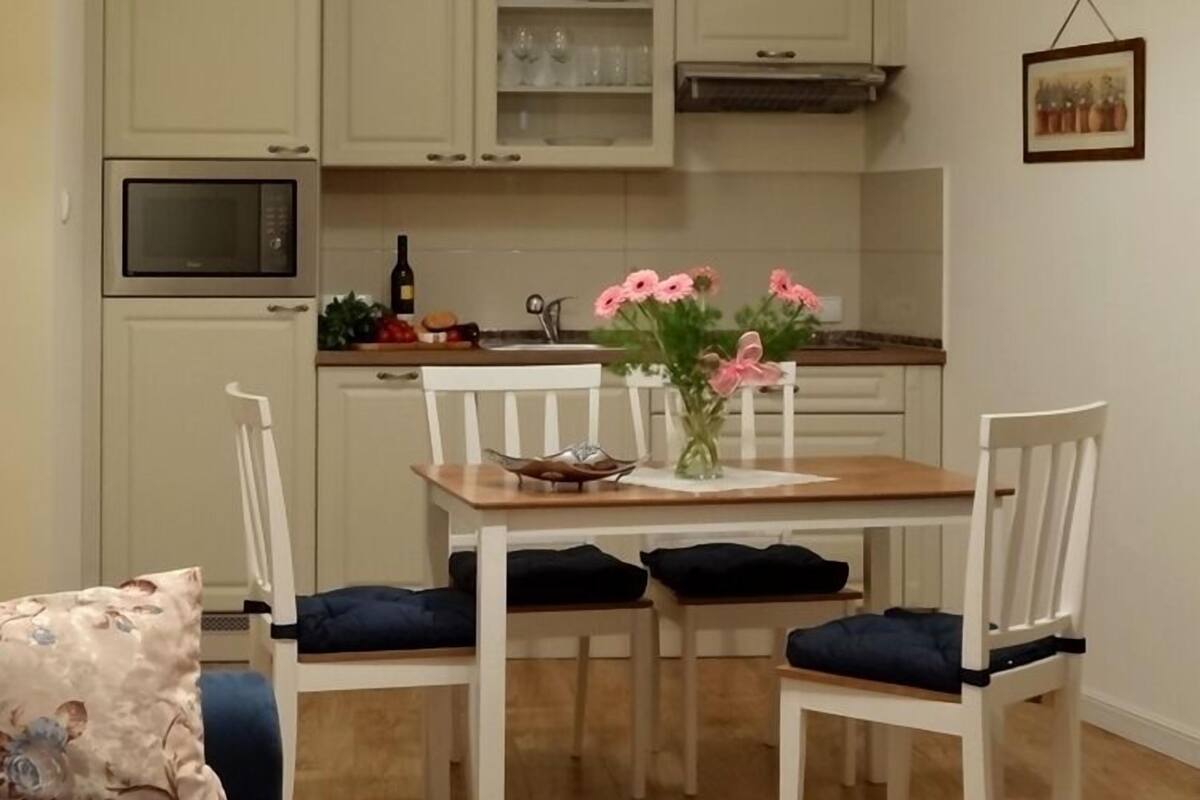 Een compacte keuken met witte kastjes, een magnetron en een eethoek met een houten tafel en stoelen.