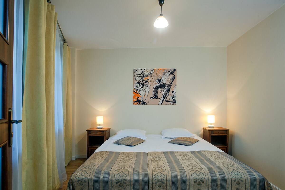 Een slaapkamer met een bed, twee nachtkastjes, een aan de muur hangend schilderij en een raam met gordijnen.