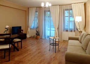 Un salon comprenant un canapé, une table à manger avec des chaises, un téléviseur, une petite table avec une lampe et de grandes fenêtres avec des rideaux.
