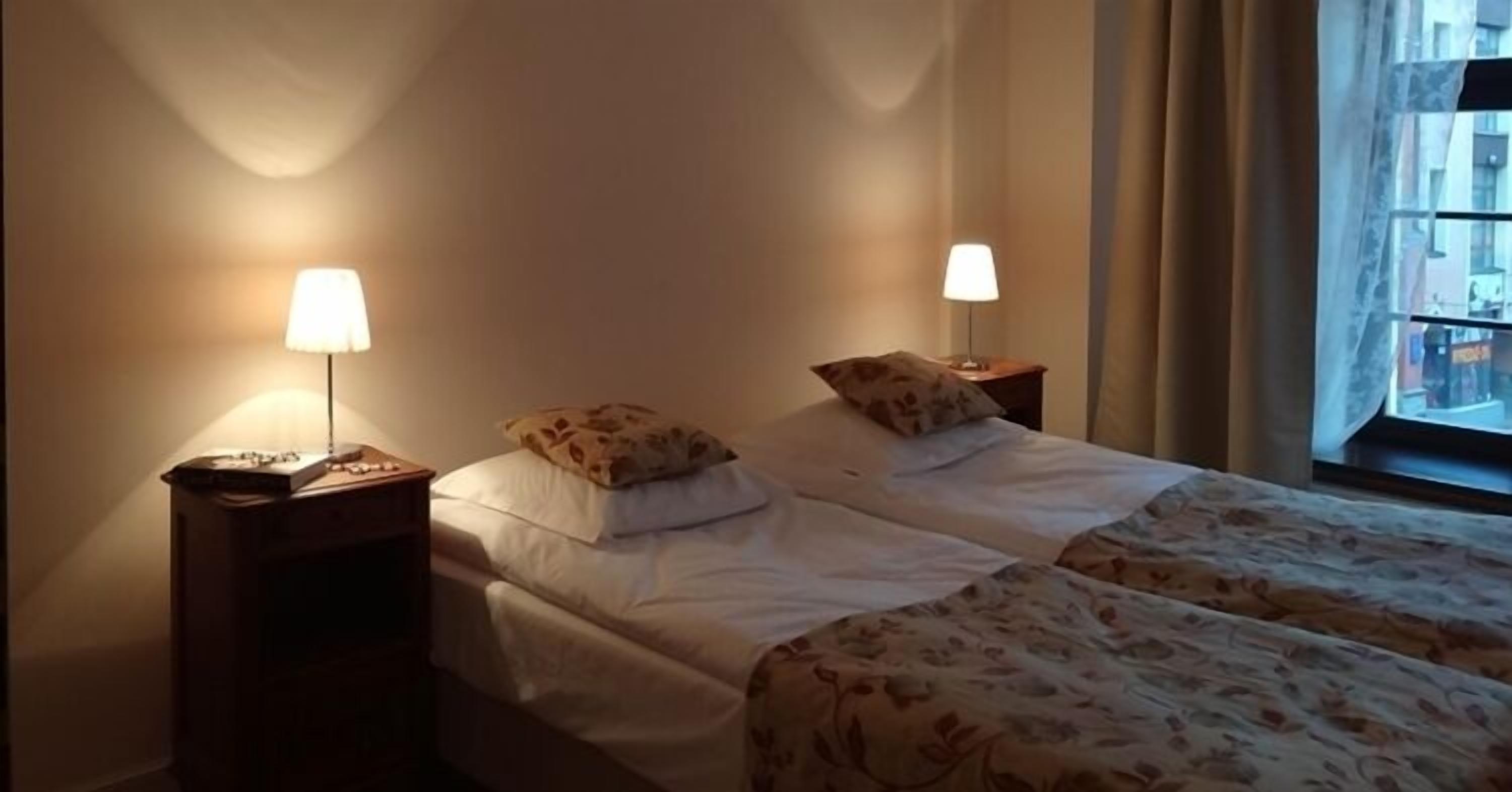 Une chambre d’hôtel avec deux lits, chacun muni d’une table de chevet et d’une lampe. La chambre est dotée d’une fenêtre avec des rideaux, et les murs sont de couleur unie.