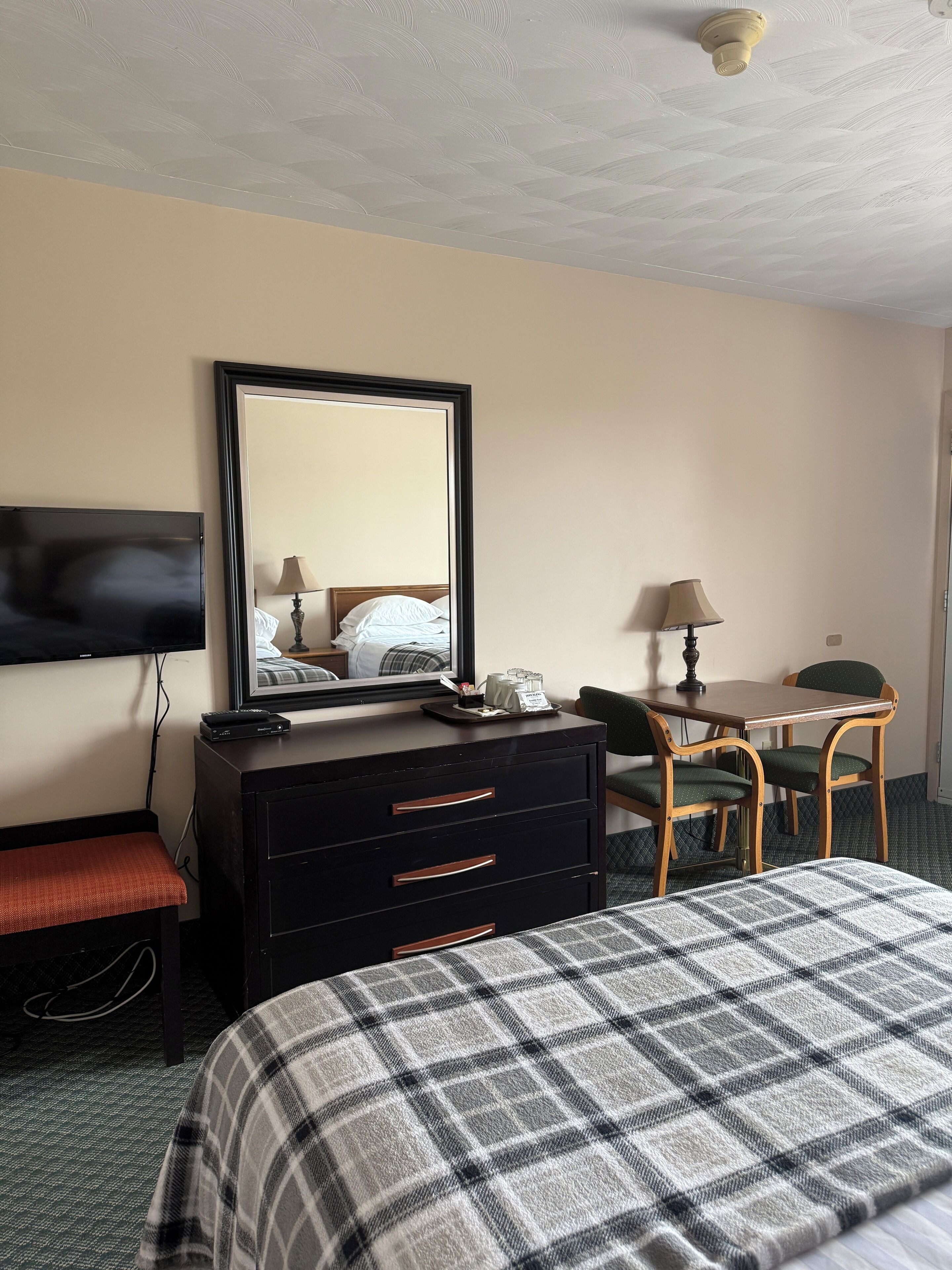 A hotel room with a bed, a dresser, a desk, a chair, a television mounted on the wall, and a ceiling fan.