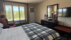 Une chambre d’hôtel avec un grand lit, un bureau, une chaise, une télévision et une fenêtre donnant sur des arbres et une route.