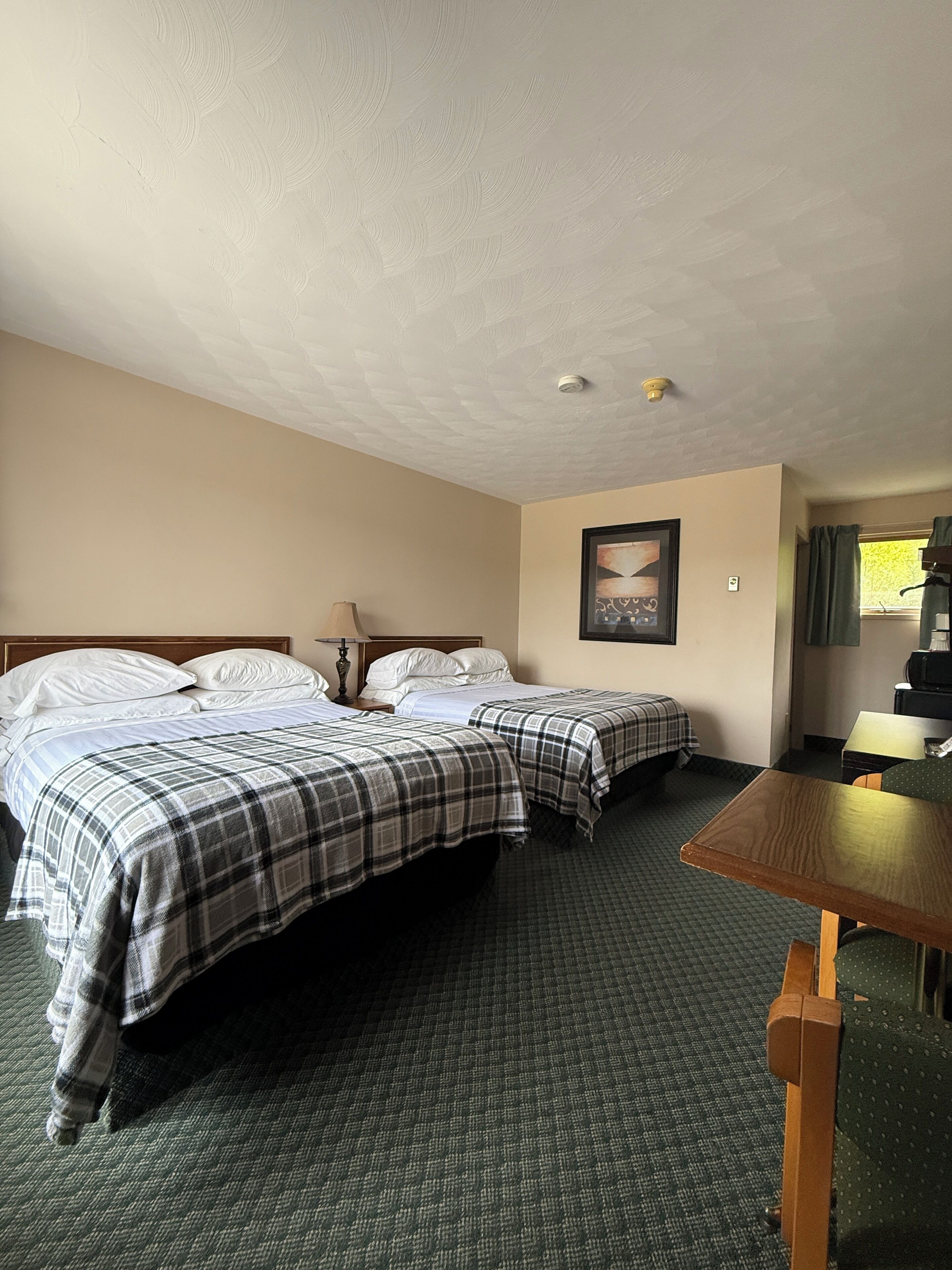 Habitación de hotel con dos camas, una mesita, una silla, un cuadro en la pared y una ventana con cortinas.