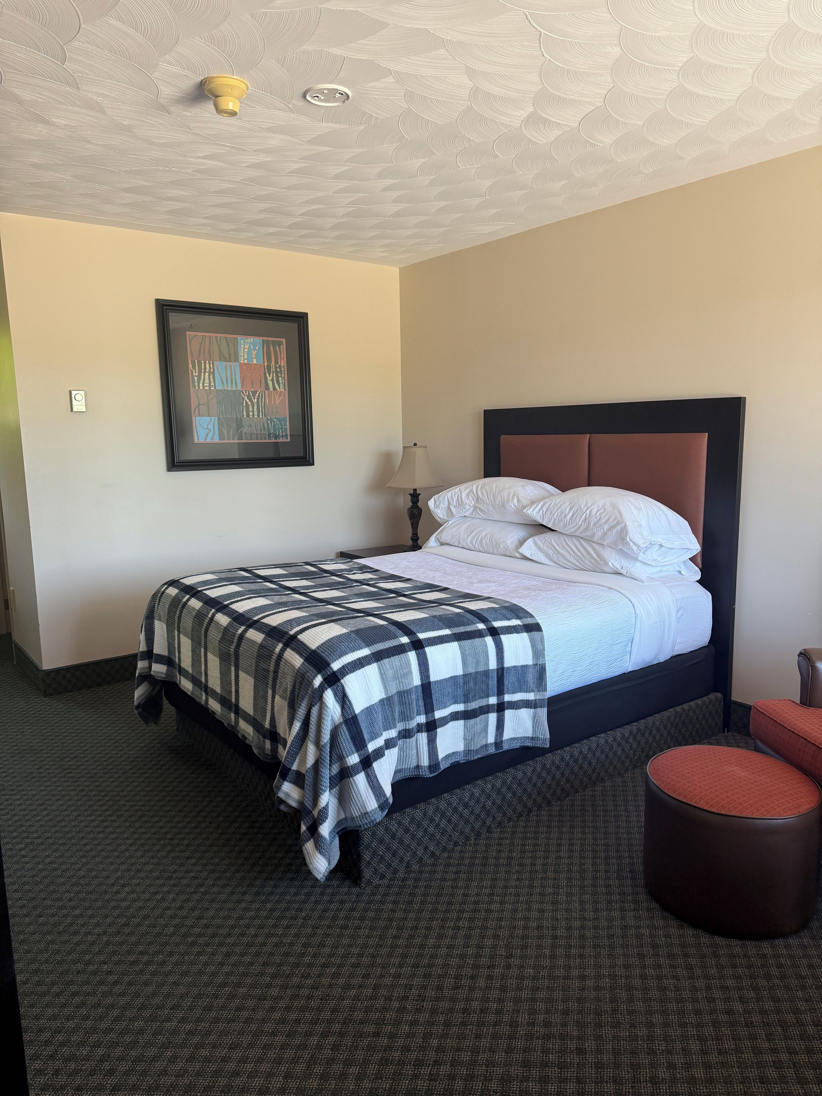 A neatly made bed with a plaid bedspread, a framed artwork on the wall, a bedside lamp, and a round ottoman.