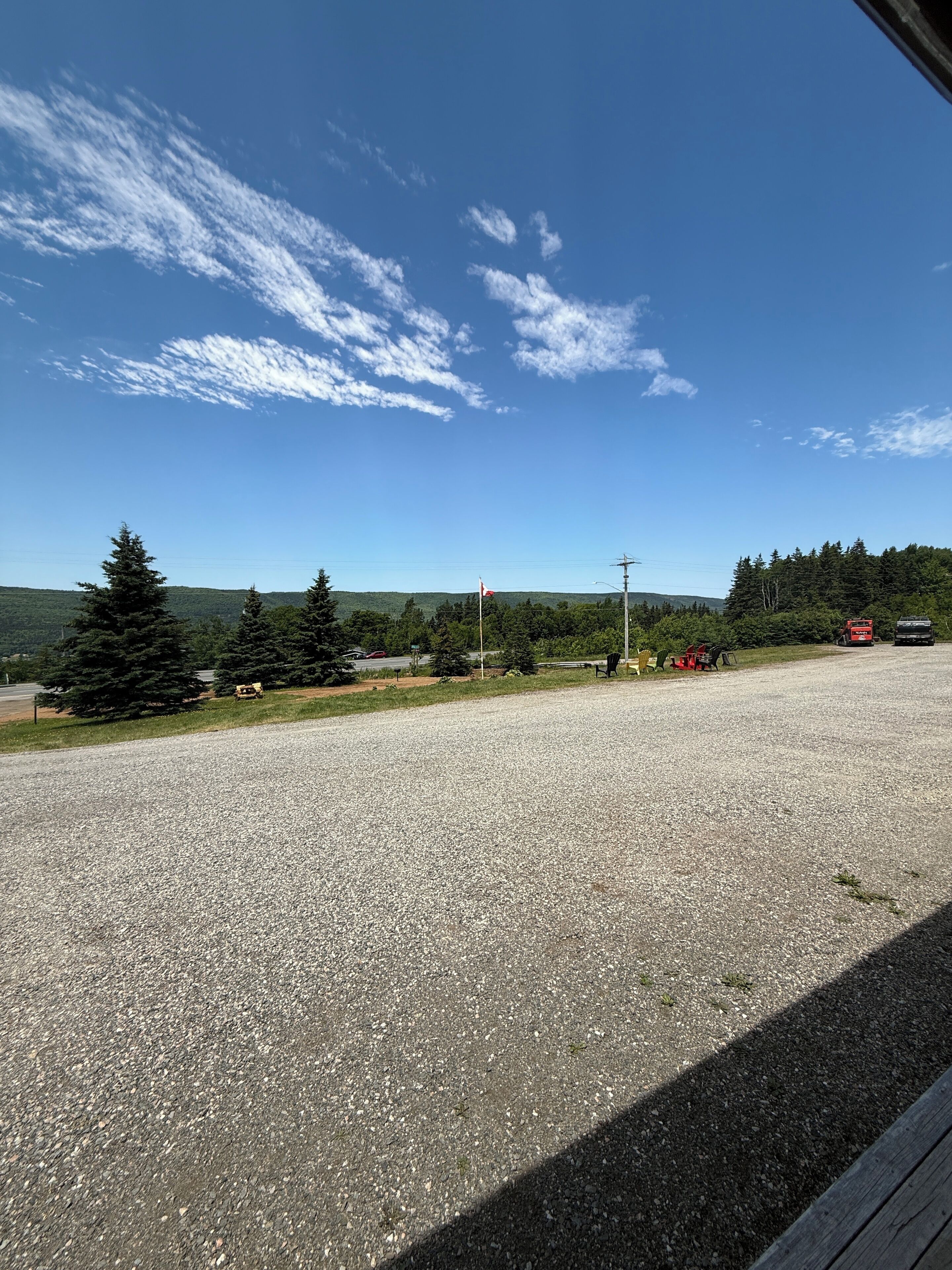 Un espace de stationnement recouvert de gravier, avec un ciel dégagé, des arbres et une vue lointaine sur une zone boisée.