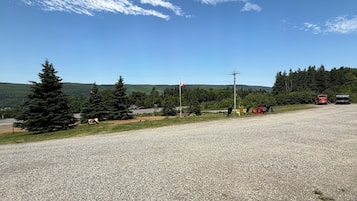 Um estacionamento de cascalho com céu claro, árvores e uma vista distante de área arborizada.