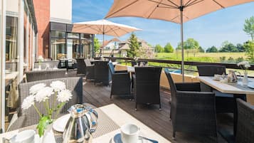 Eine Terrasse im Freien mit Korbmöbeln, einem Tisch mit weißer Tasse und Untertasse sowie einem großen Sonnenschirm, der Schatten spendet.
