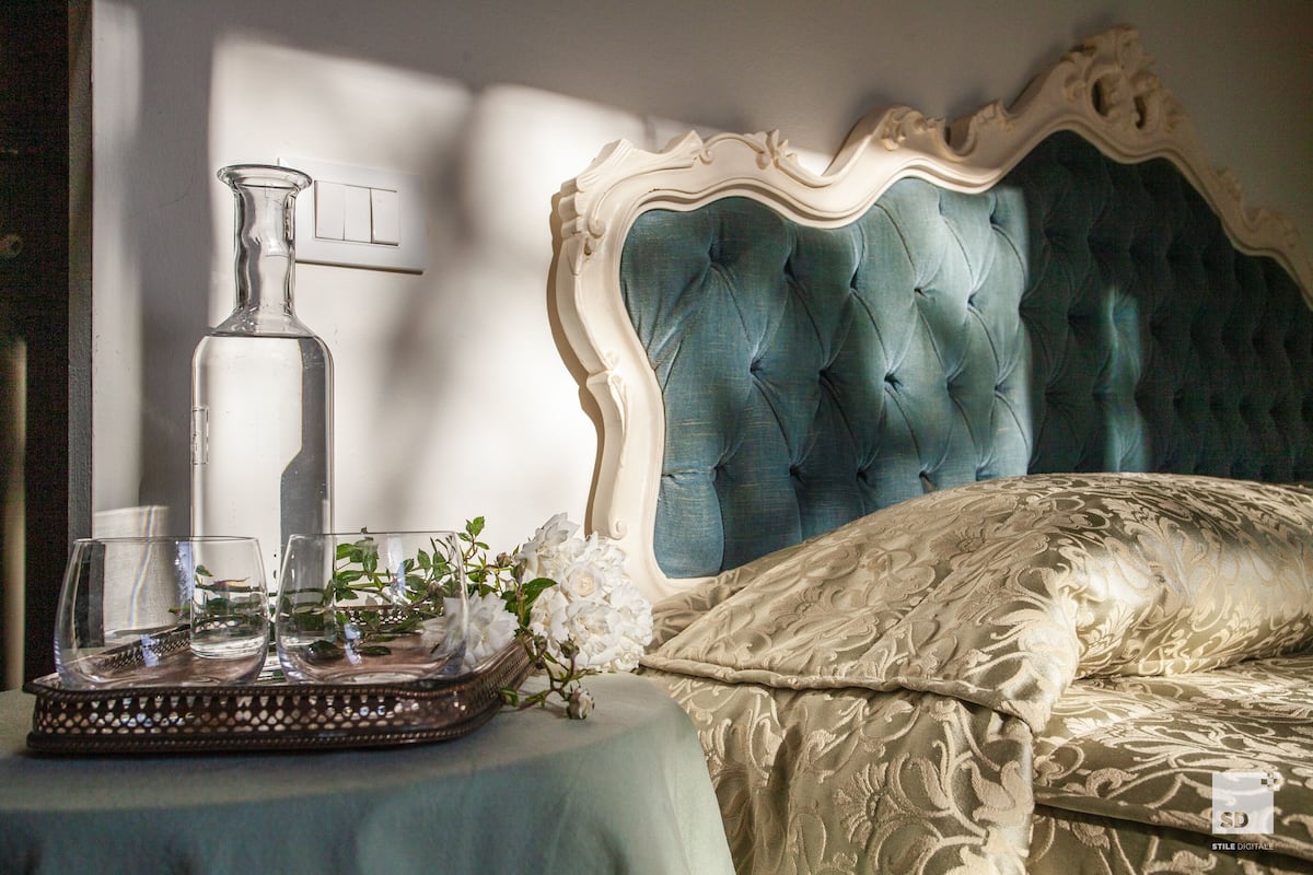 Een bed met een gestoffeerd hoofdbord, een nachtkastje met een dienblad waarop glazen en een vaas staan, en een lichtschakelaar aan de muur.