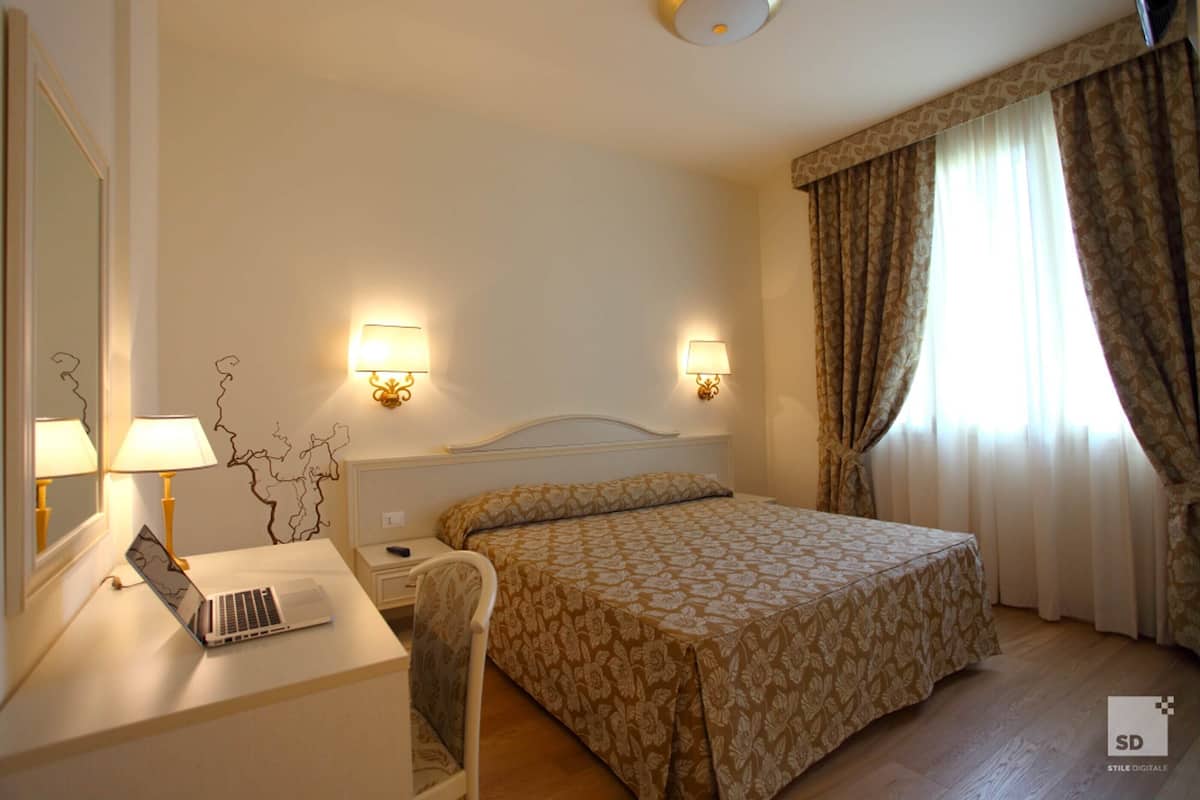 Hotelkamer met een bed, een bureau met een laptop, twee bedlampen en een raam met gordijnen.