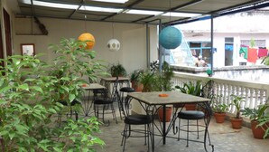An outdoor patio area with tables and chairs, surrounded by potted plants and hanging lanterns.