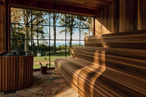 Una sauna con suelo de piedra, un banco de madera y una ventana que da a árboles y una masa de agua.