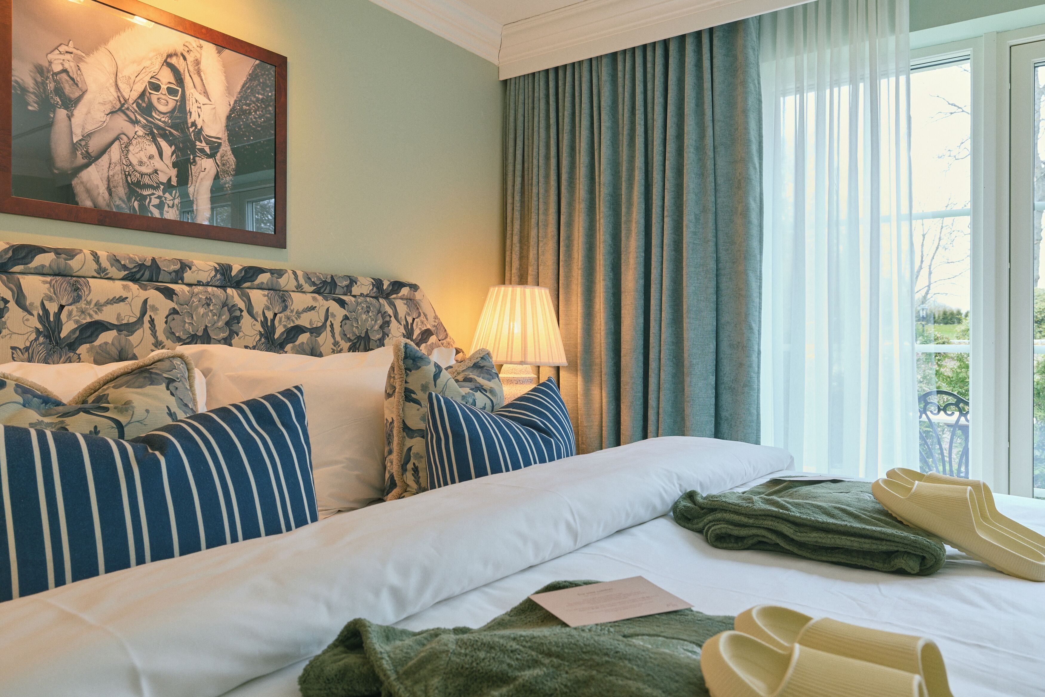A hotel room with a bed, pillows, a lamp, a window with curtains, and a framed picture on the wall.