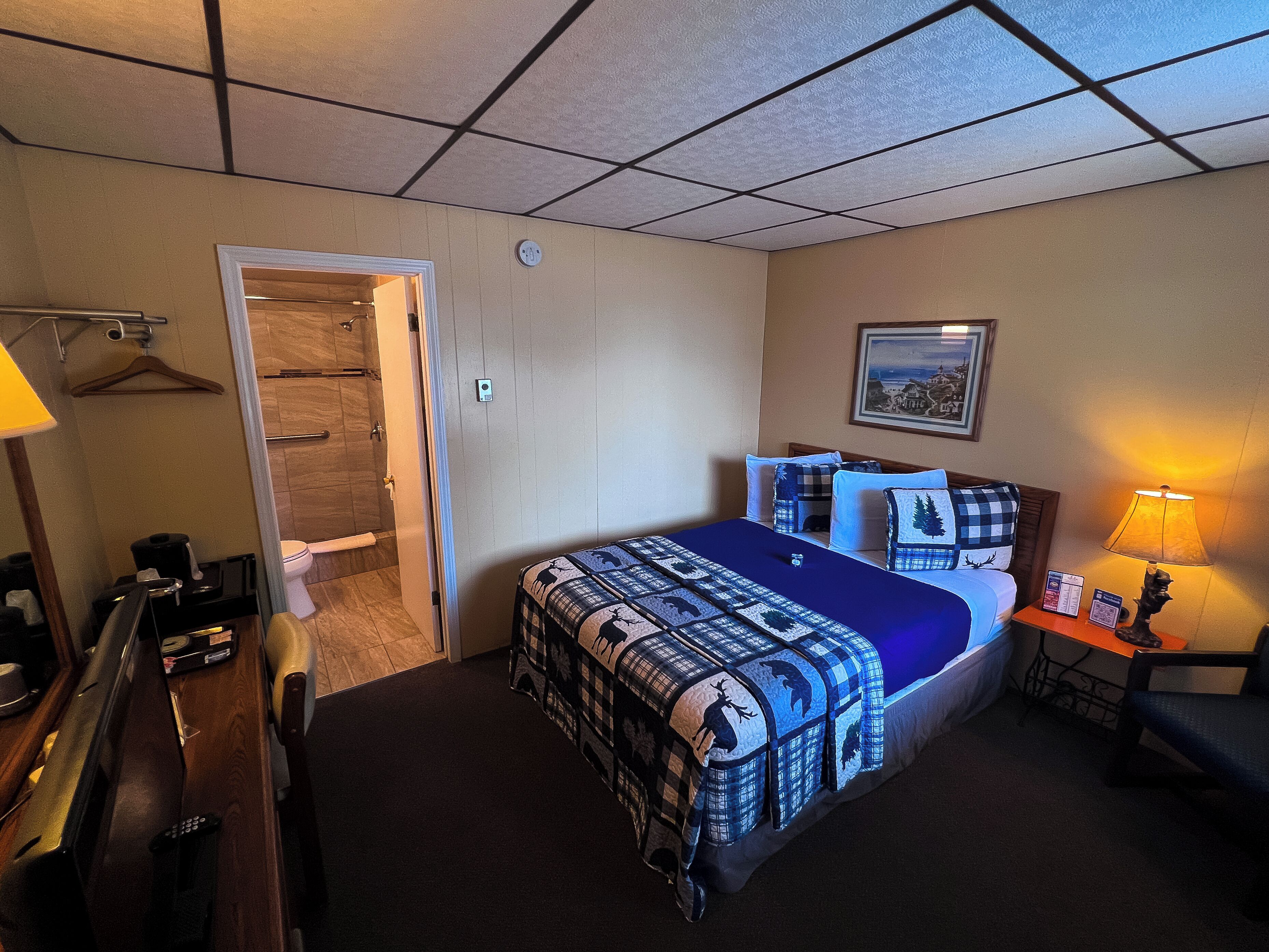 A hotel room with a bed, a desk, a chair, a lamp, a painting on the wall, and a door leading to a bathroom.