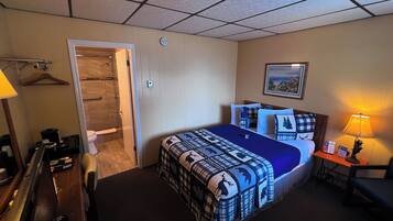 A hotel room with a bed, a desk, a chair, a lamp, a painting on the wall, and a door leading to a bathroom.
