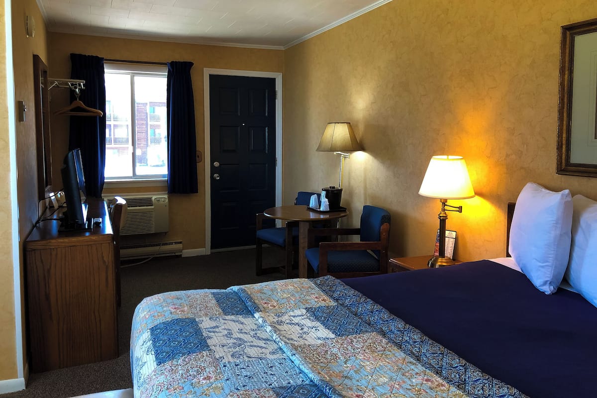 A hotel room with a bed, a desk with a computer, a chair, a lamp, and a window with curtains.