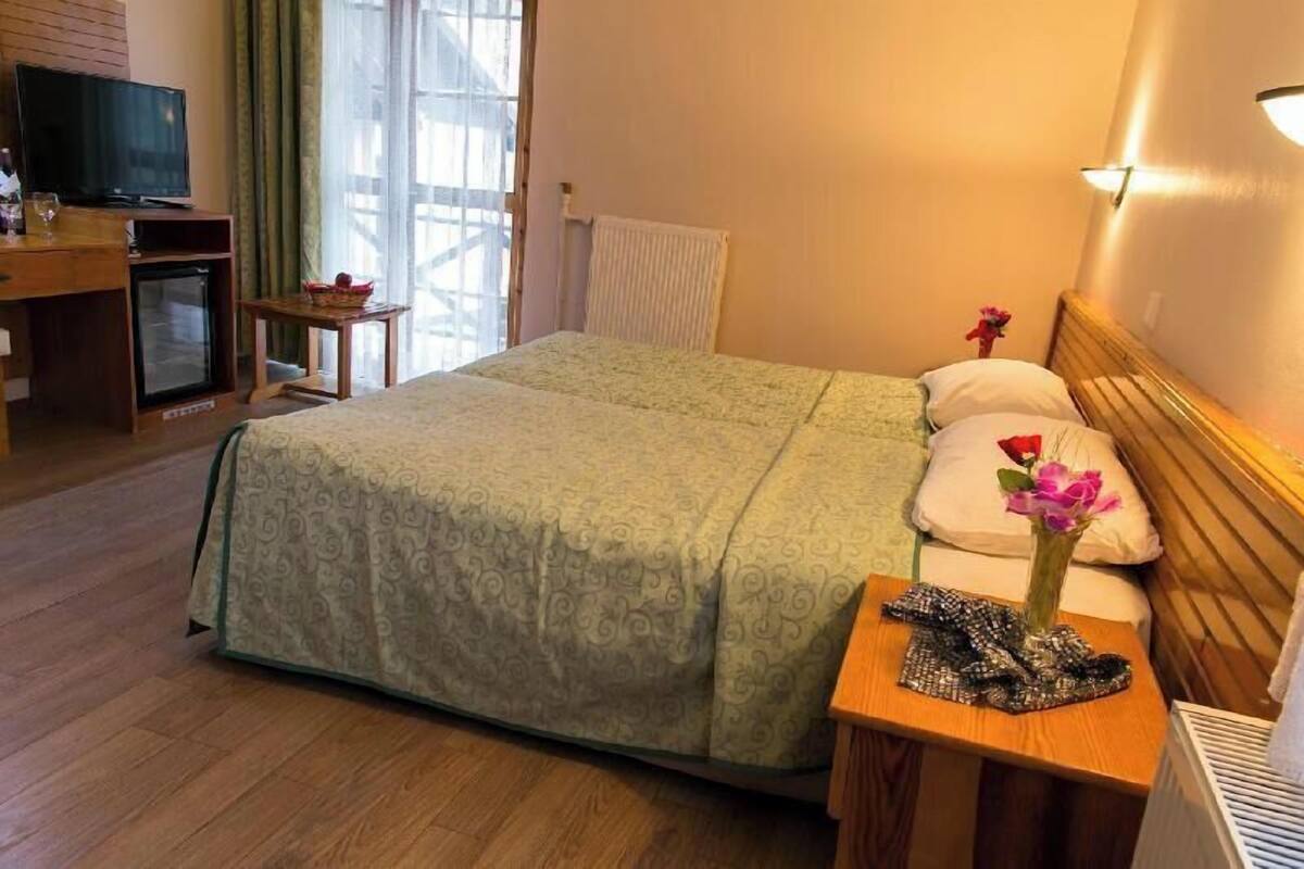 A hotel room with a bed, a small table with a vase of flowers, a television, a desk with a computer, and a window with curtains.