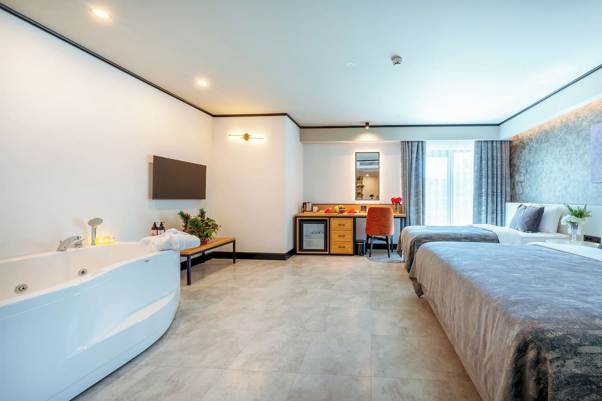 A modern hotel room with a large bed, a separate desk area, a bathtub, and a large window.
