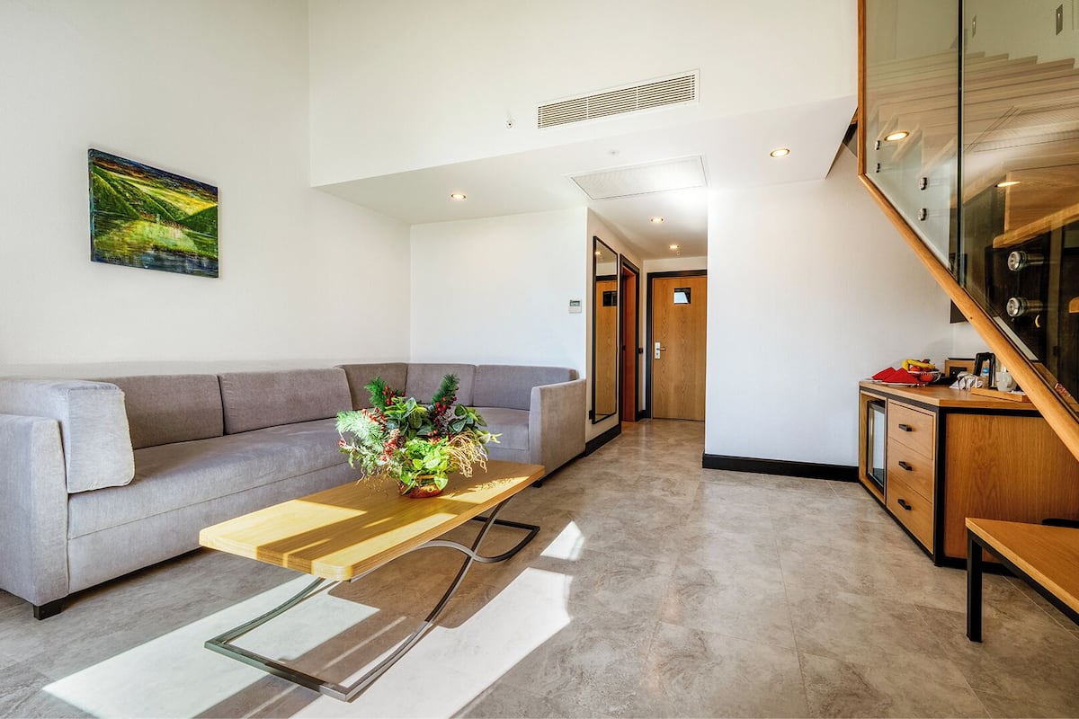 A modern living room with a grey sofa, a wooden coffee table with a plant, and a glass coffee table. There is a staircase to the right and a door leading to another room.