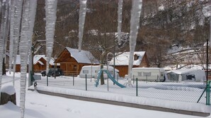 A snow-covered outdoor area with a wooden cabin, a playground, and a fenced parking area. - Camping Prado Verde (Vilamos)