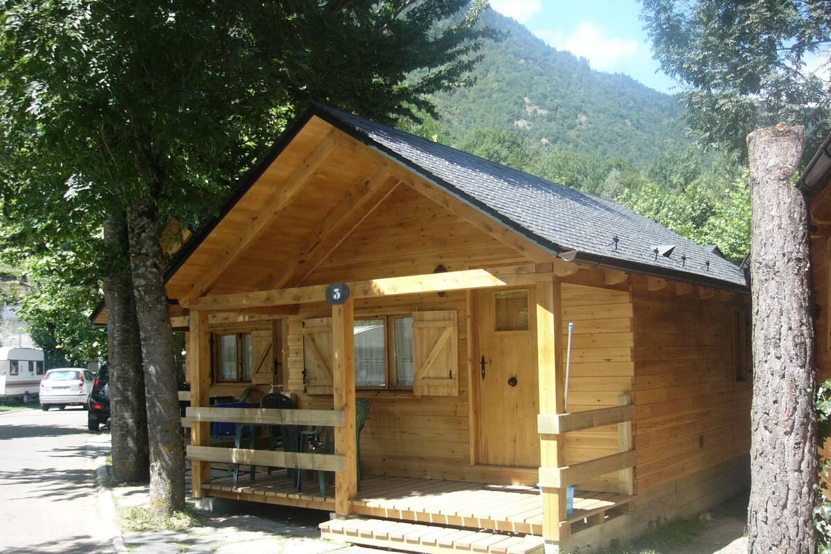 Una cabaña de madera con un porche, una pequeña terraza y montañas al fondo.