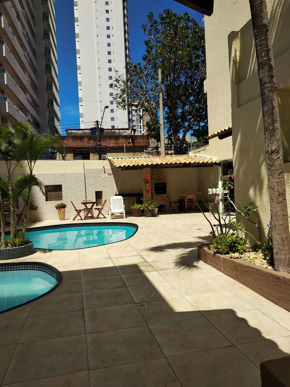 Una zona de piscina con suelo de baldosas, un rincón con sillas para sentarse y un edificio con techo de tejas al fondo.