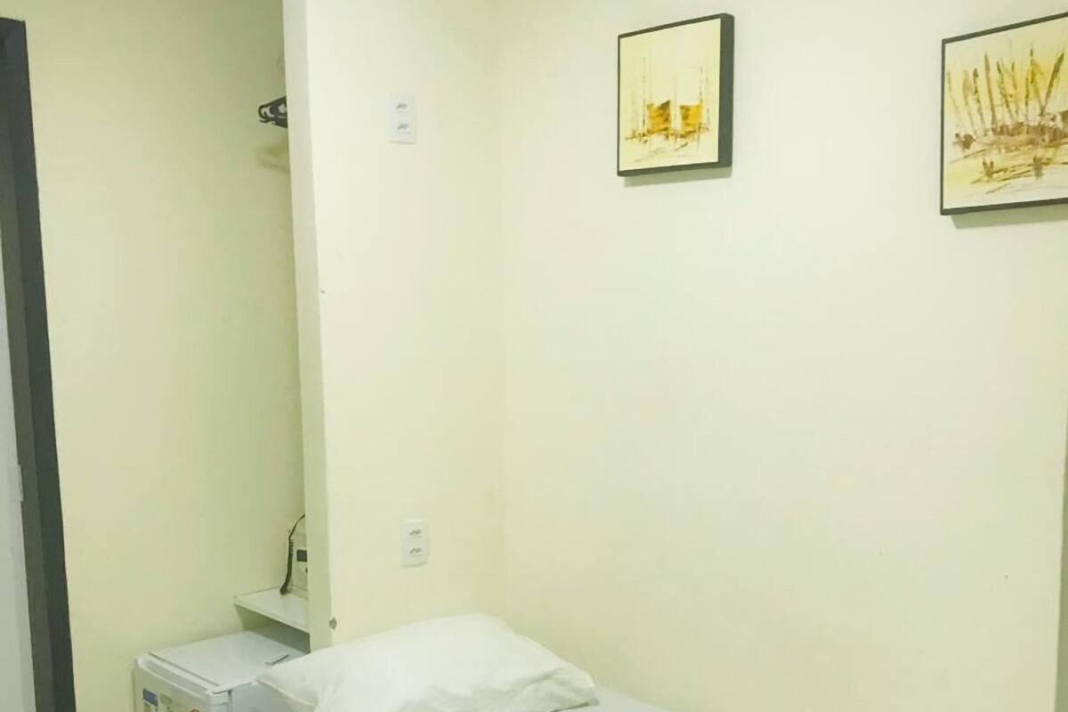 A single-bed room with a white bed, a small heater, and two framed artworks on the wall.