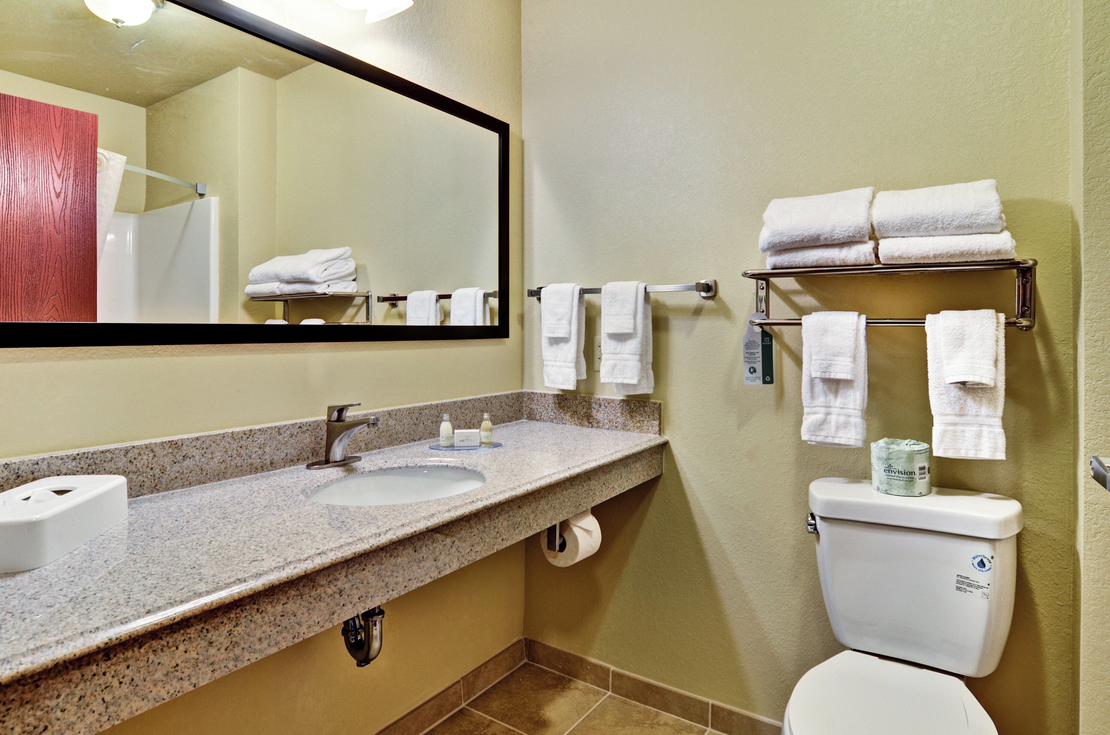 A bathroom with a granite countertop sink, a mirror, a toilet, and a towel rack with folded towels.