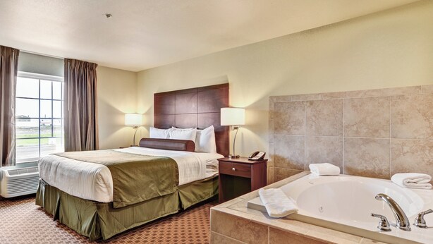Habitación de hotel con cama, mesita de noche con lámpara, baño con bañera y ventana con cortinas.