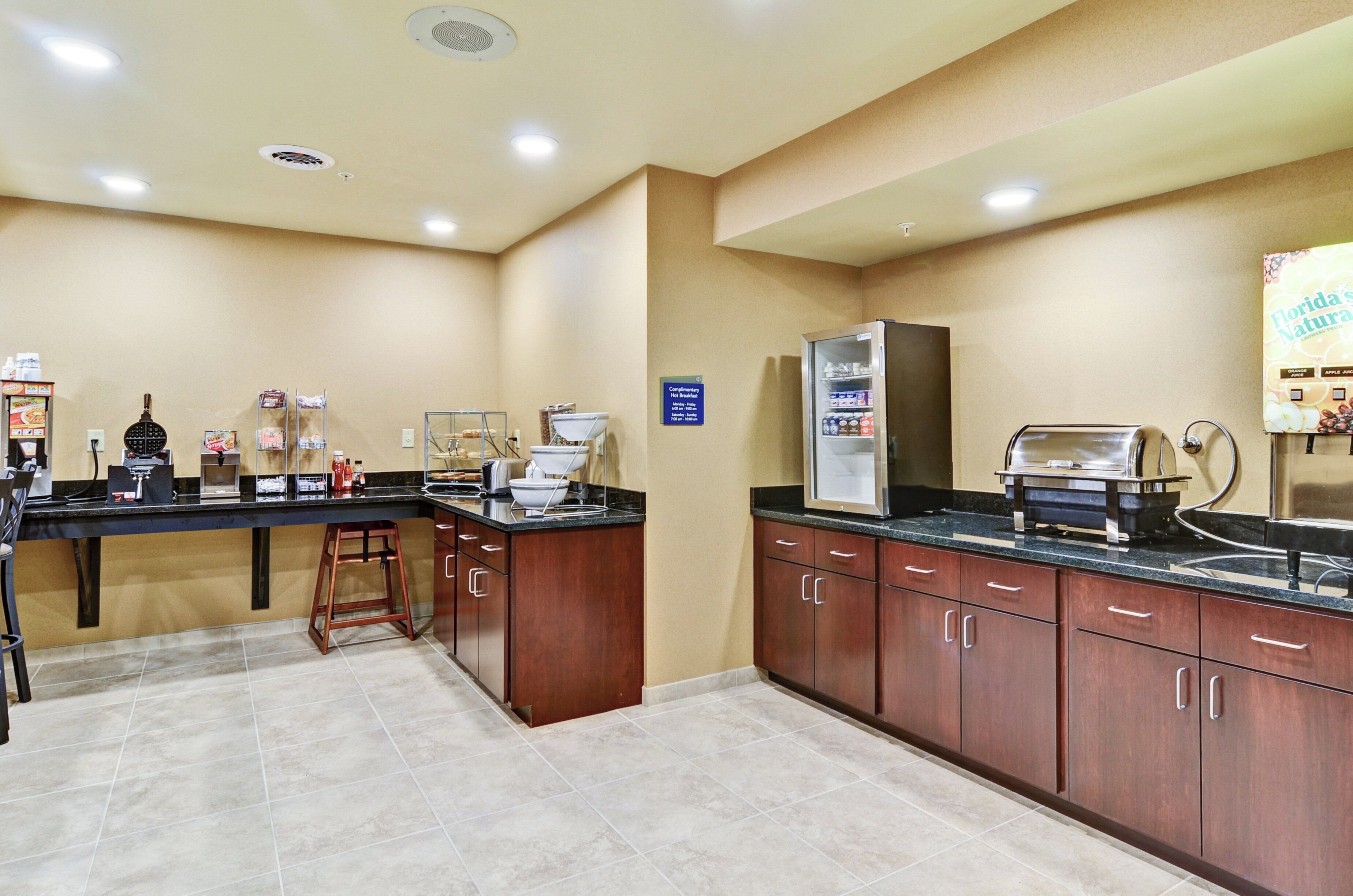 Cocina de hotel con estación de café, refrigerador y un letrero que indica desayuno de cortesía.