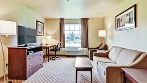 Habitación de hotel con televisión de pantalla plana, cómoda de madera, sofá, mesita, escritorio con silla y ventana con cortinas.