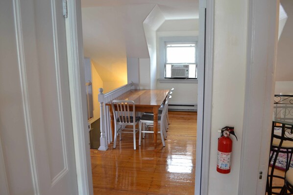 A dining area with a wooden table and chairs, a fire extinguisher on the wall, and a window with an air conditioning unit. - Allston Red House (Boston)