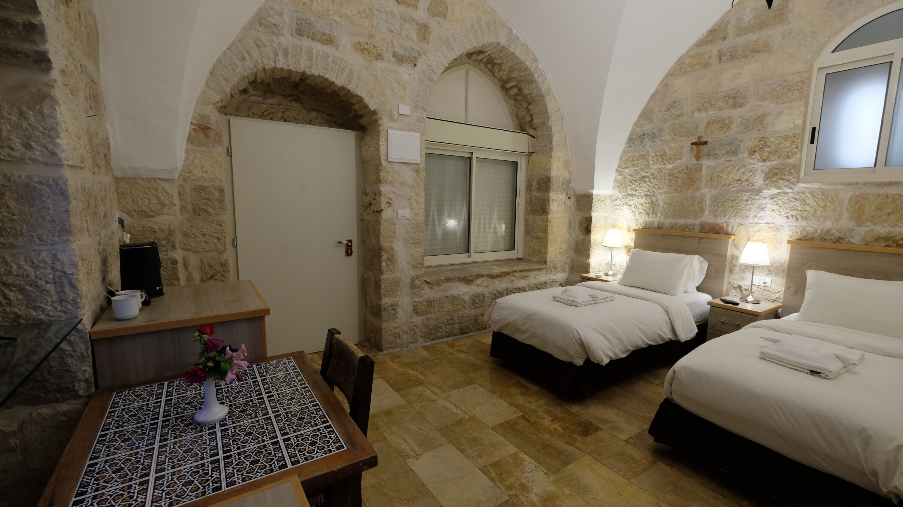 A room with two beds, a small table with a vase of flowers, and a stone wall.