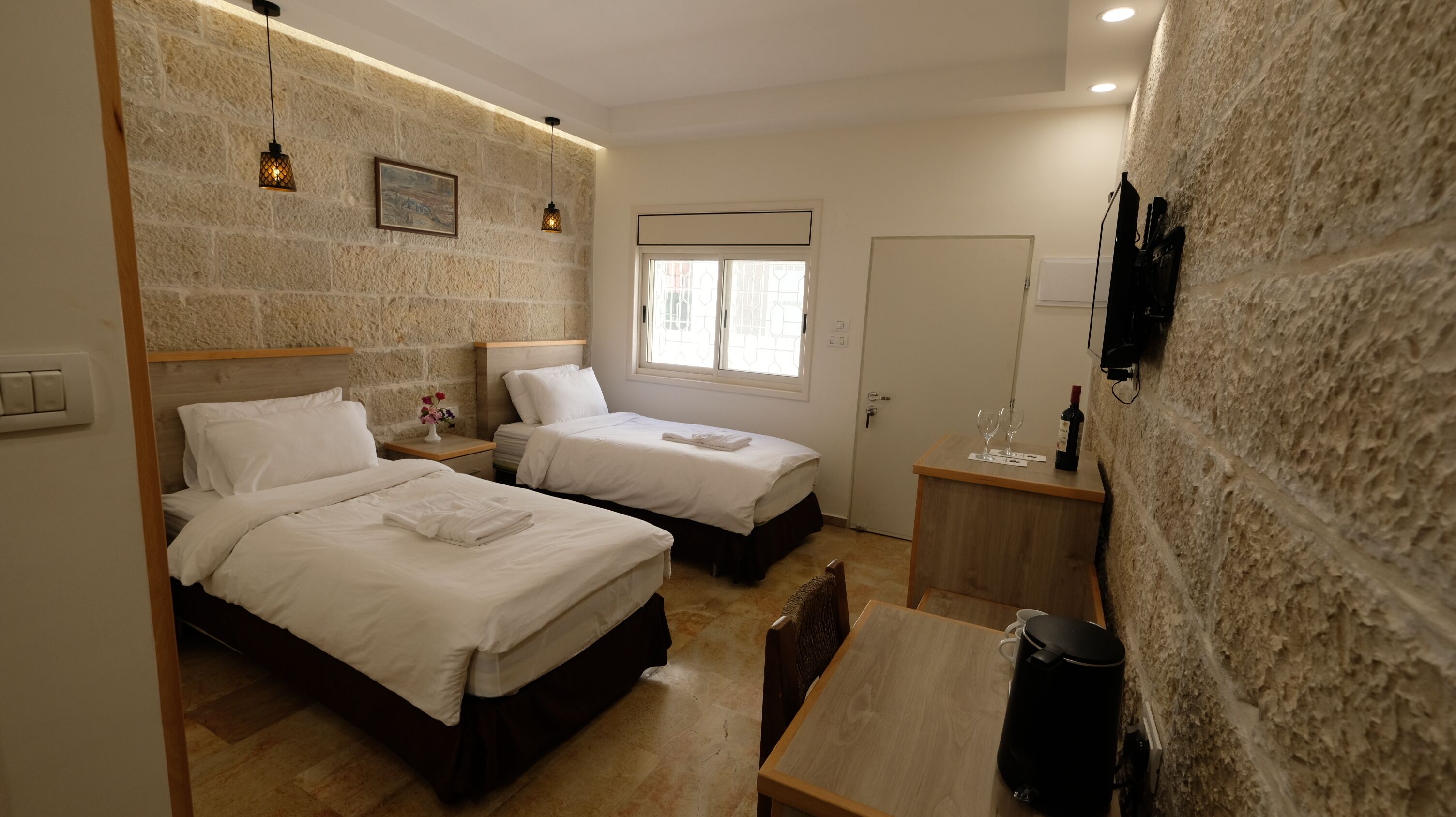 A hotel room with two beds, a wooden table, a coffee maker, a wine bottle, and a stone wall.