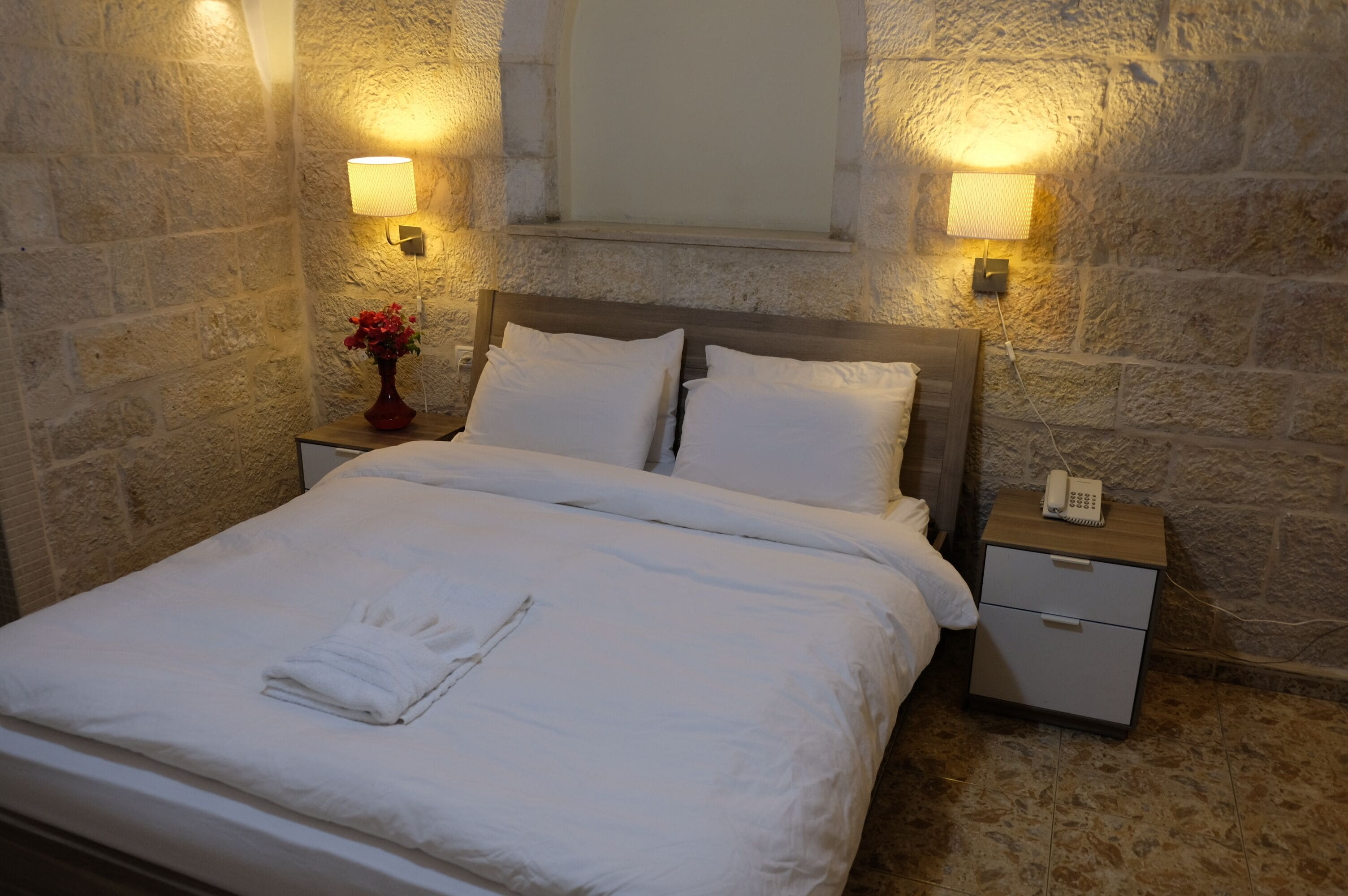 A neatly made bed with white linens and a folded towel, two bedside lamps, a nightstand with a phone, and a vase with flowers.
