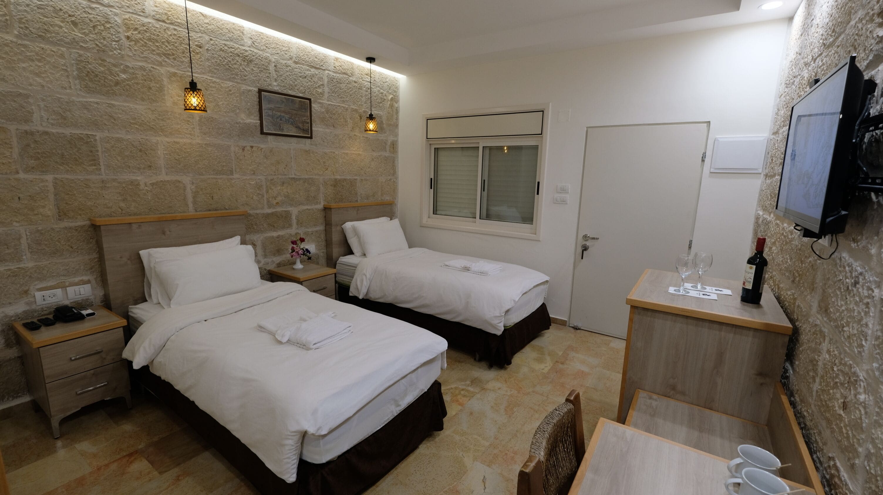 A hotel room with two beds, a wooden nightstand, a desk with a chair, a television, and a stone wall.