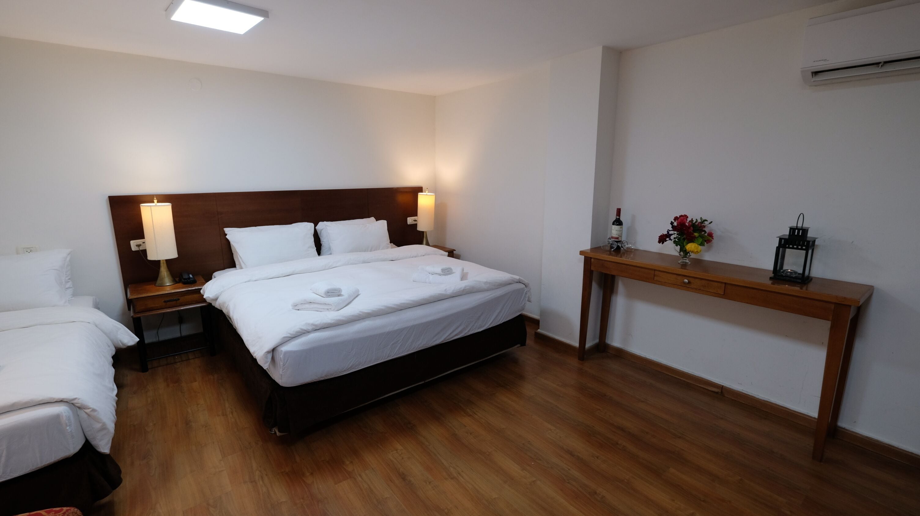 A hotel room with a large bed, two bedside tables with lamps, a wooden nightstand, a wooden desk, and a small table with a bottle and flowers.