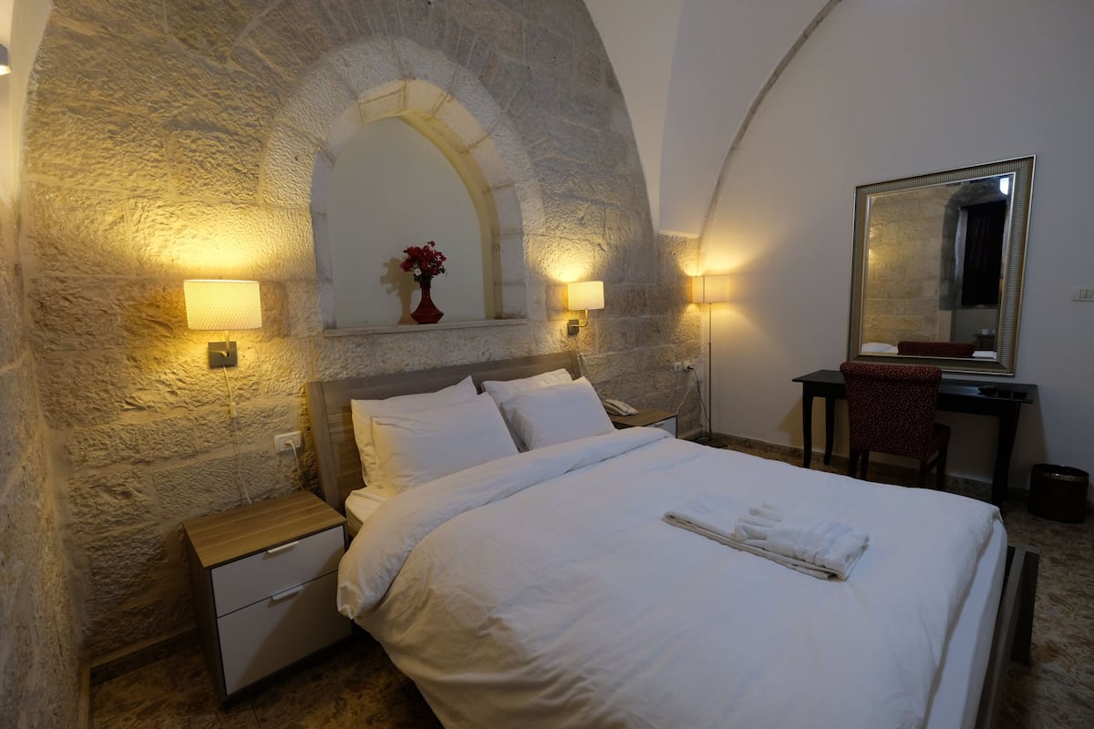 A bedroom with a stone wall, a bed with white linens, a nightstand, a desk with a chair, and a mirror.