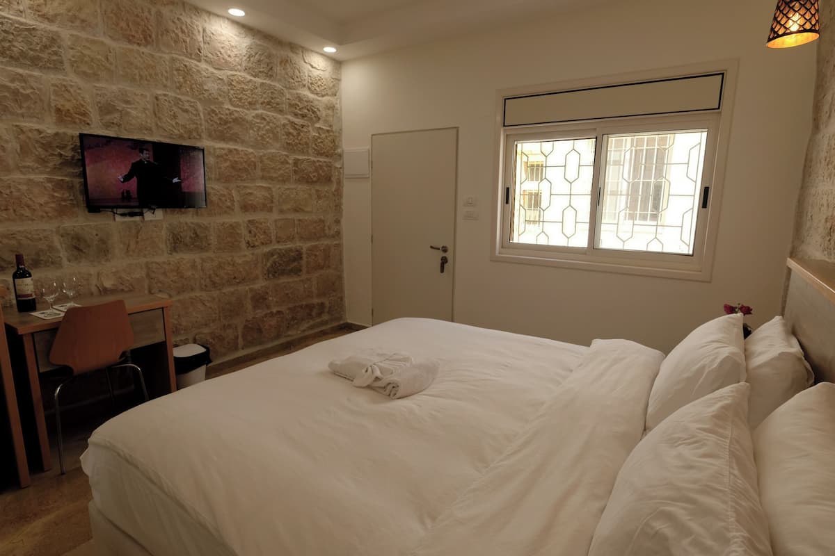 A hotel room with a stone wall, a bed with white linens, a desk with a chair, a television, and a window with a geometric pattern.