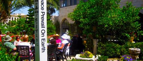 Un patio con un letrero que dice "May Peace Prevail On Earth", rodeado de exuberante vegetación y personas cenando.