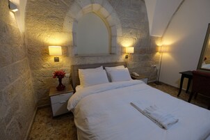 Un dormitorio con paredes de piedra, una cama con ropa blanca, una mesita de noche con un florero y dos lámparas fijadas a la pared.