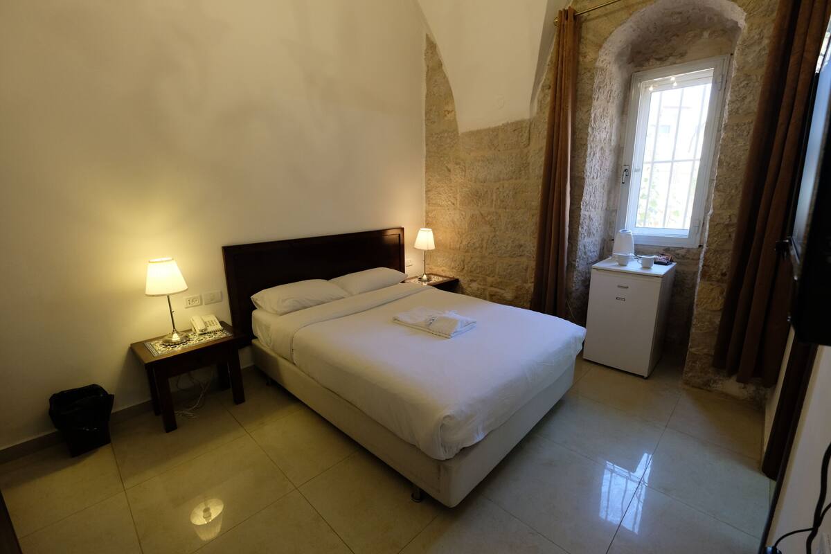 A hotel room with a bed, bedside lamps, a small desk, a refrigerator, and a window with curtains.