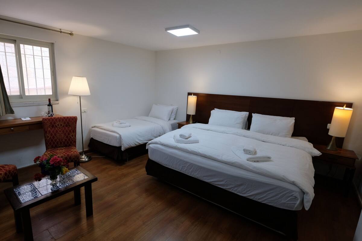 A hotel room with two single beds, a large bed, a desk, a chair, a table with a vase of flowers, and a window with curtains.