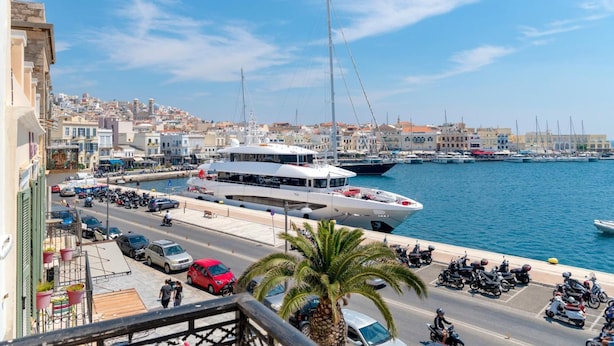 A marina with boats, a road with vehicles, and buildings in the background. - Florence (Syros)